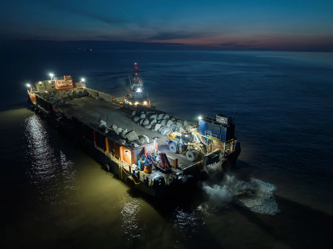 A sea barge at night carries a delivery of Armourstone granite boulders from Glensanda to Blue Anchor Beach, Somerset.