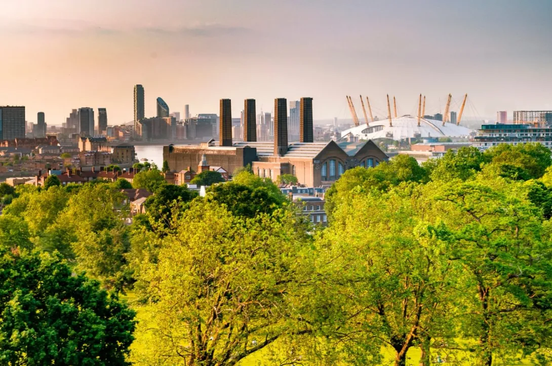 An image of the London skyline with trees in the foreground and buildings including the O2 and Canary Wharf in the background